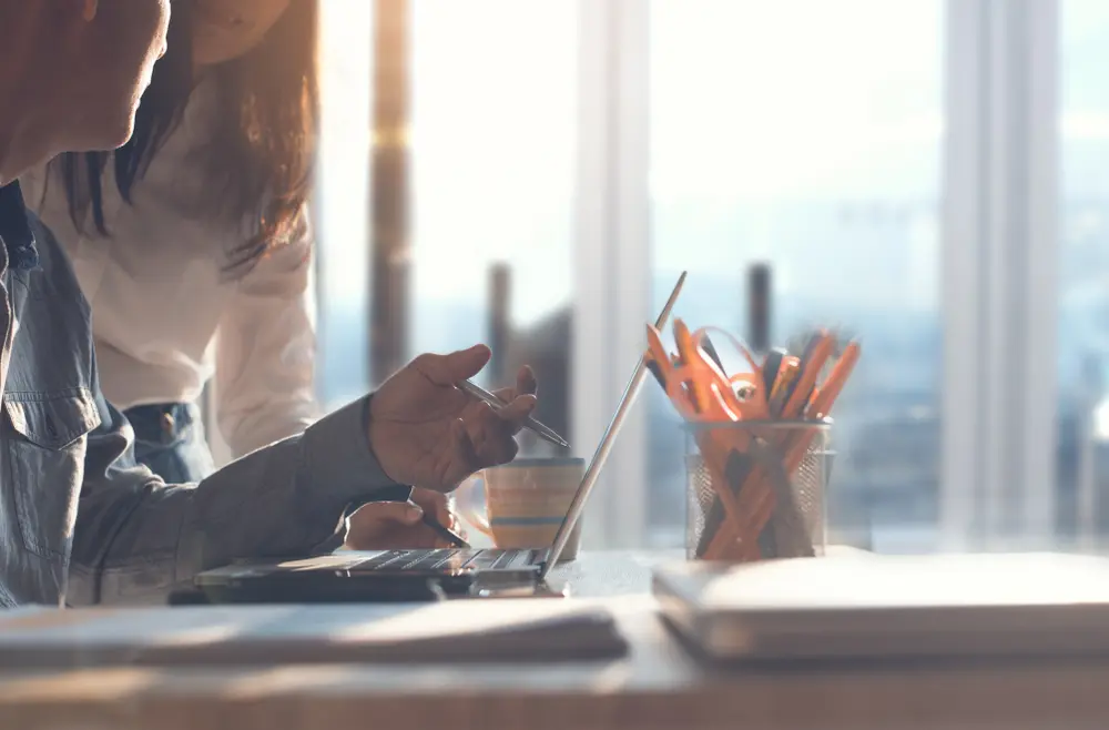 A person uses a pen to point at a laptop screen while collaborating with a colleague in a bright, sunlit office space with a pencil holder and coffee mug on the desk.