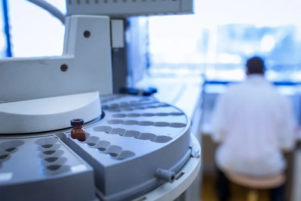 A small amber vial is positioned in an automated laboratory carousel, with a blurred scientist in a white lab coat working in the background of a modern, sterile medical or research facility.