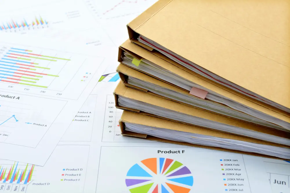 A stack of brown manila folders, some with colorful adhesive tabs, sits atop a variety of business reports featuring pie charts and bar graphs.