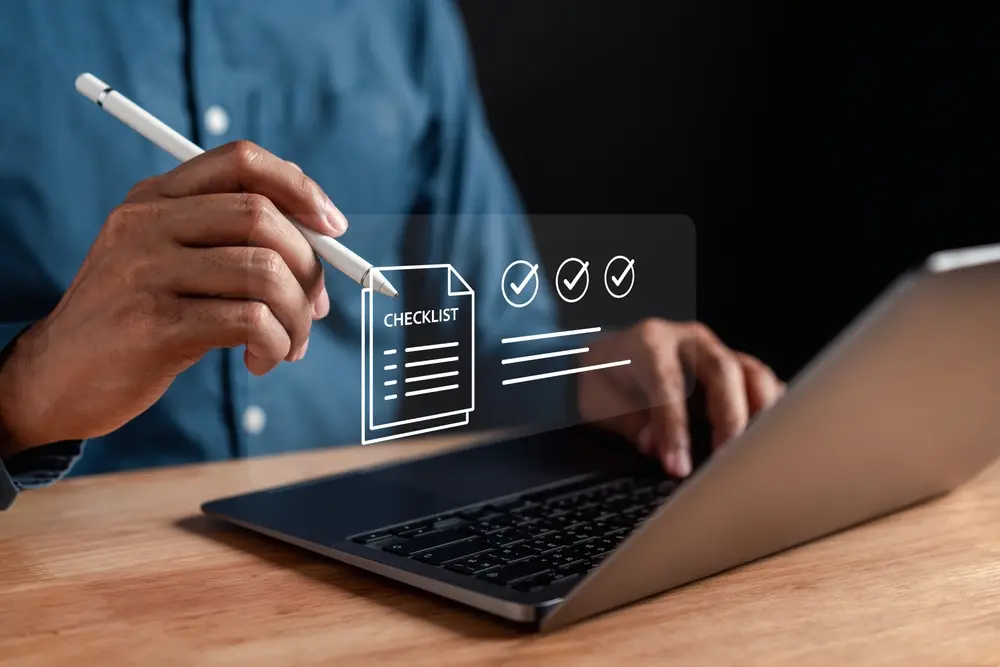 A person using a stylus and a laptop with a translucent digital "CHECKLIST" icon overlay featuring three completed checkmarks.