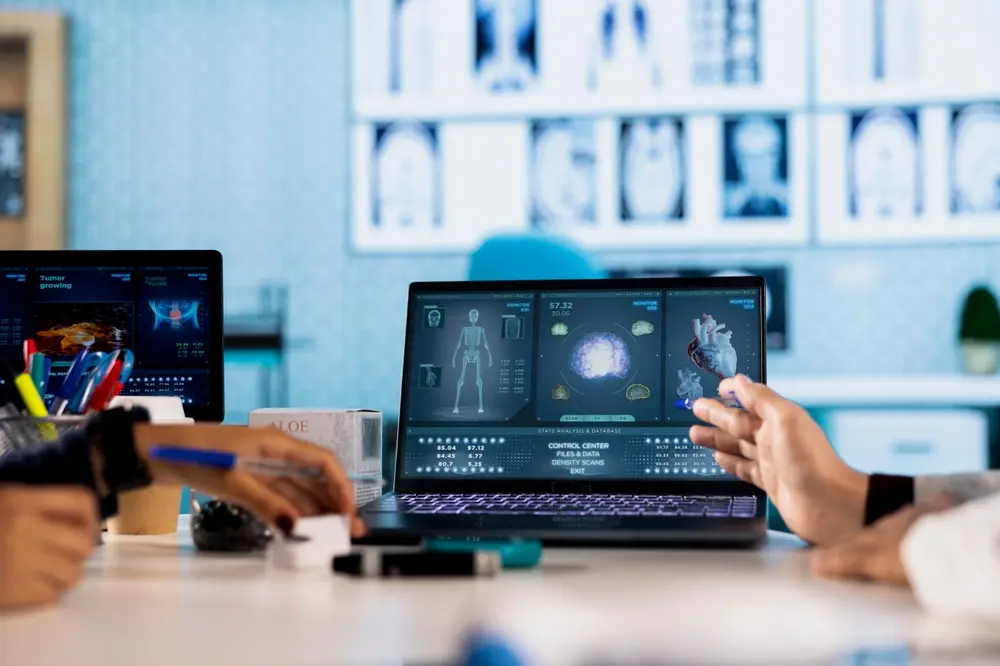 Healthcare professionals discussing medical data displayed on a laptop screen, showing anatomical scans of a skeleton, brain, and heart in a modern clinical setting.
