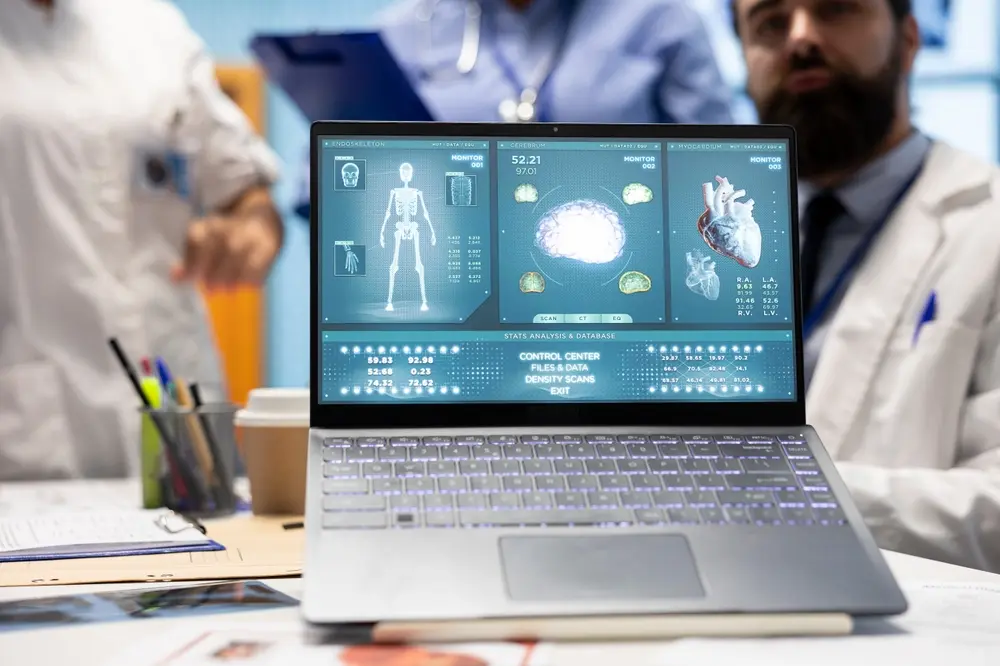 A laptop screen displaying detailed medical data visualizations of a human skeleton, brain, and heart, with healthcare professionals in lab coats blurred in the background.