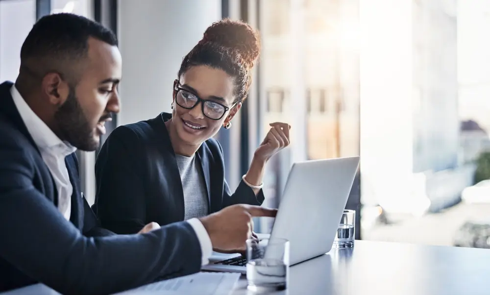 Two professionally dressed colleagues, a man and a woman, smiling and collaborating while looking at a laptop screen in a bright office setting.
