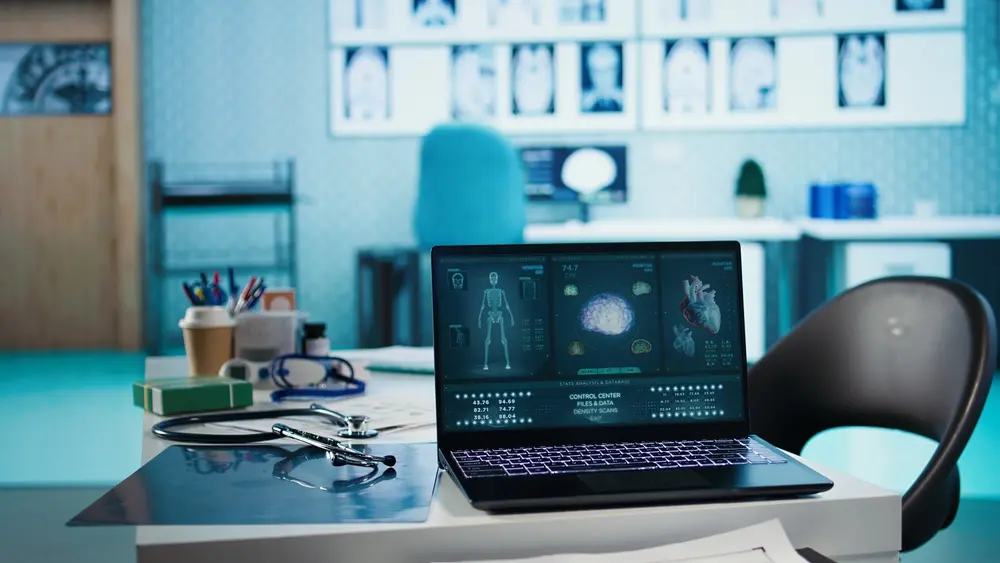 A laptop on a white desk displaying medical interface scans of a skeleton, brain, and heart, situated in a brightly lit medical office with a stethoscope and X-rays in the foreground.