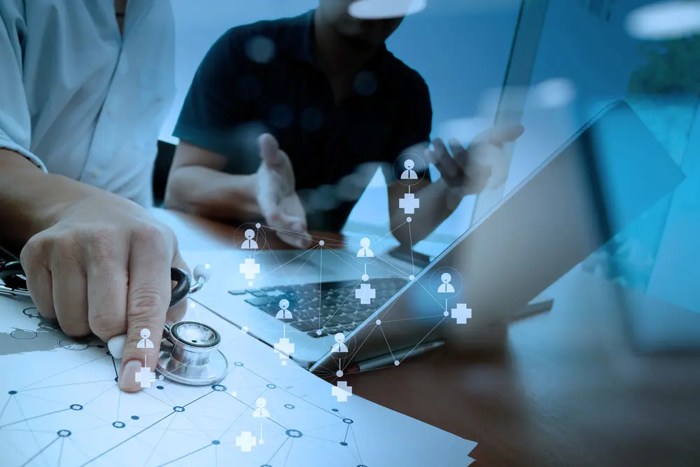 A healthcare professional holding a stethoscope points at a document while collaborating with a colleague at a laptop, with a glowing digital network overlay of medical crosses and people icons connecting the elements of the scene.