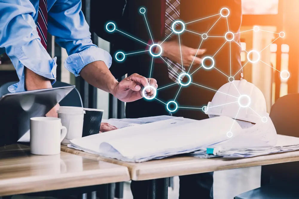 Two men in business attire collaborating over blueprints and a white hard hat on a desk, with a glowing digital network graphic overlaid on the scene.